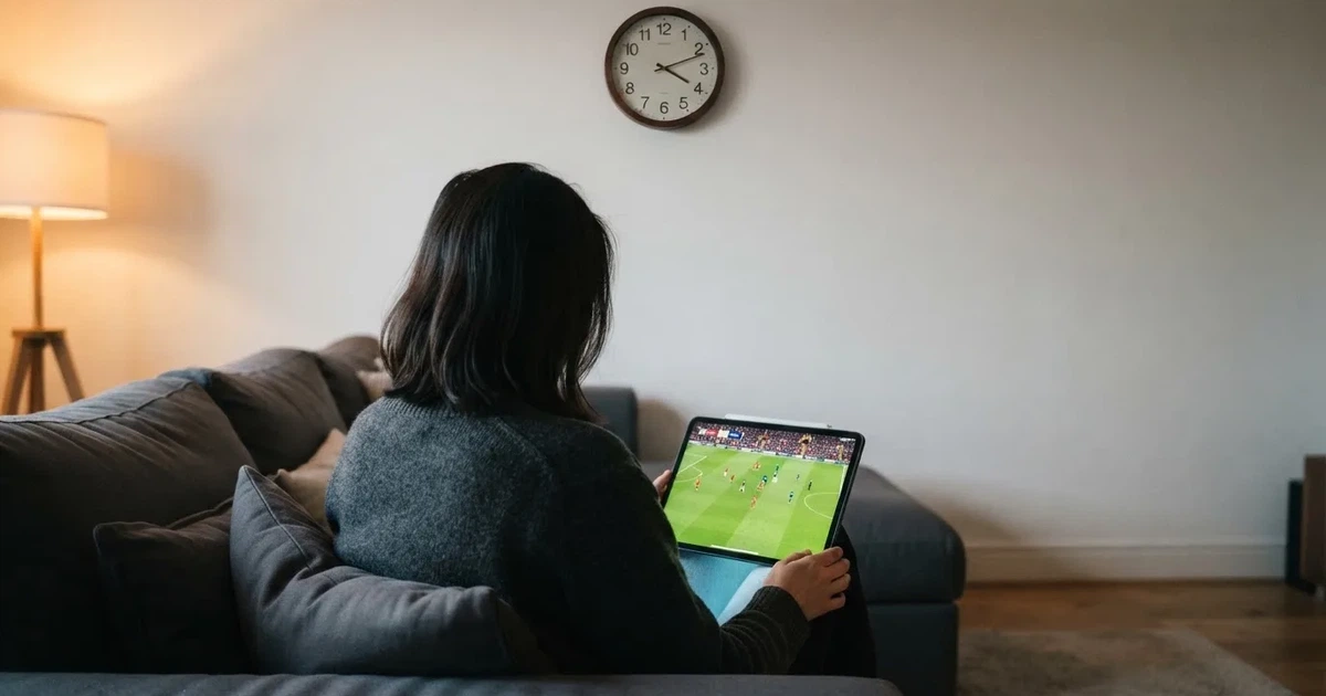 Een persoon op de bank kijkt peinzend naar een tablet, op de achtergrond een klok aan de muur